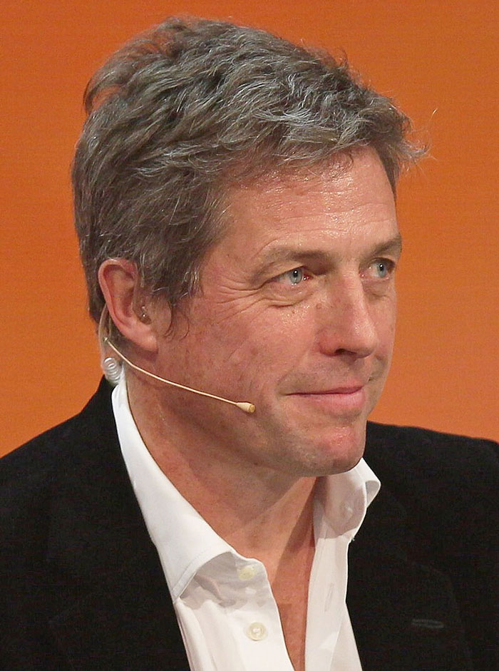 Middle-aged man wearing a headset mic, dressed in a white shirt and black jacket, speaking in a professional setting.