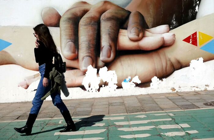 Woman walking past a mural of large hands on the street, capturing unseen, unposed moments in street photography.