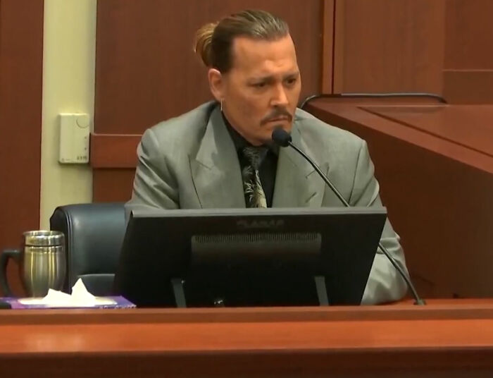 Celebrity appearing out-of-touch during courtroom testimony, wearing a gray suit and speaking into a microphone.