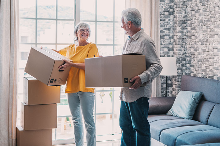 Elderly couple smiling and carrying moving boxes inside a living room, representing parents visiting their son's home.