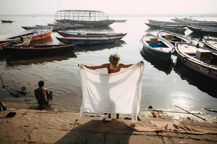 Elderly man dries white cloth by riverbank with boats in background, candid everyday moments from Asia by Gil Kreslavsky.