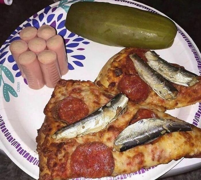 Unusual food combination with pepperoni pizza topped with sardines, sliced hot dogs, and a large pickle on a paper plate.