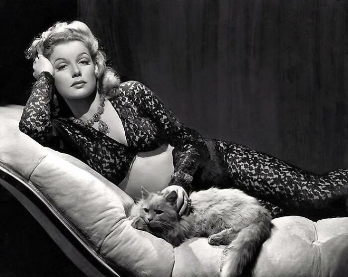 Vintage cat photo of a glamorous woman reclining on a chaise lounge, gently petting a fluffy feline companion.