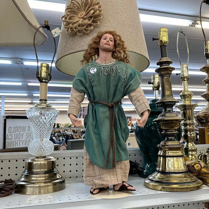 Porcelain figurine dressed in vintage clothes standing among brass and crystal lamps at a thrift store with bizarre items.