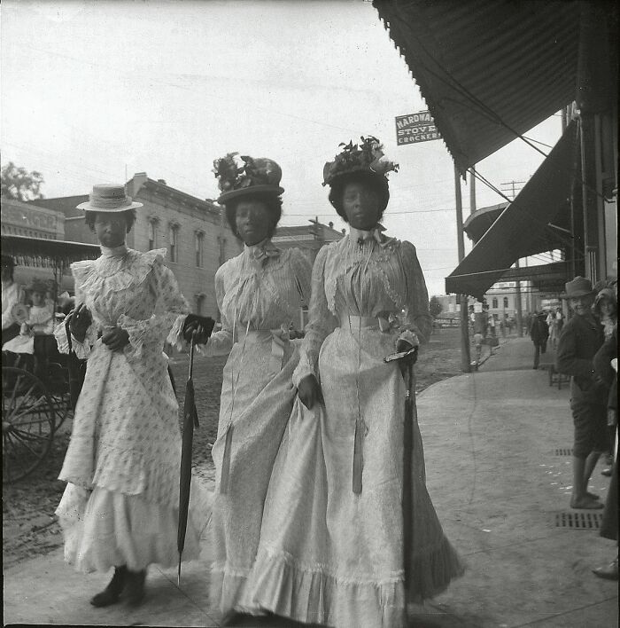 Three women in vintage dresses and hats holding parasols, walking on a historic city street in a historical photo.