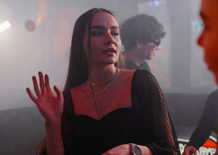 Young woman in a dimly lit party scene sharing wild and embarrassing drinking stories with friends around her.