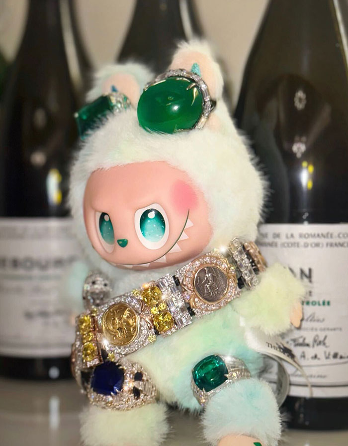 Labubu doll wearing jeweled accessories with green stones, displayed in front of wine bottles.