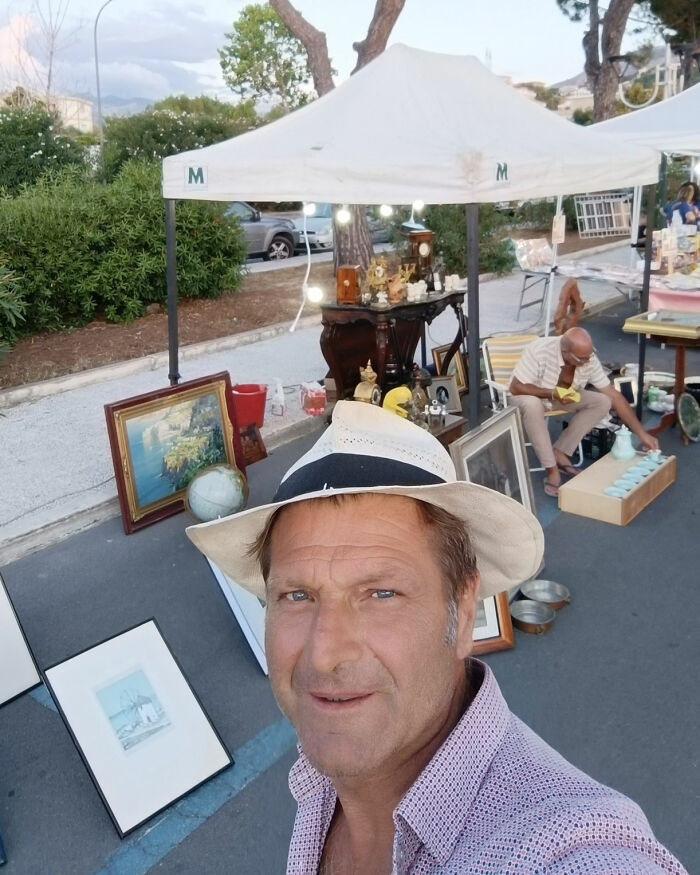 Man wearing hat taking selfie at outdoor market stall with antiques, related to toxic broccoli sandwich investigation case.
