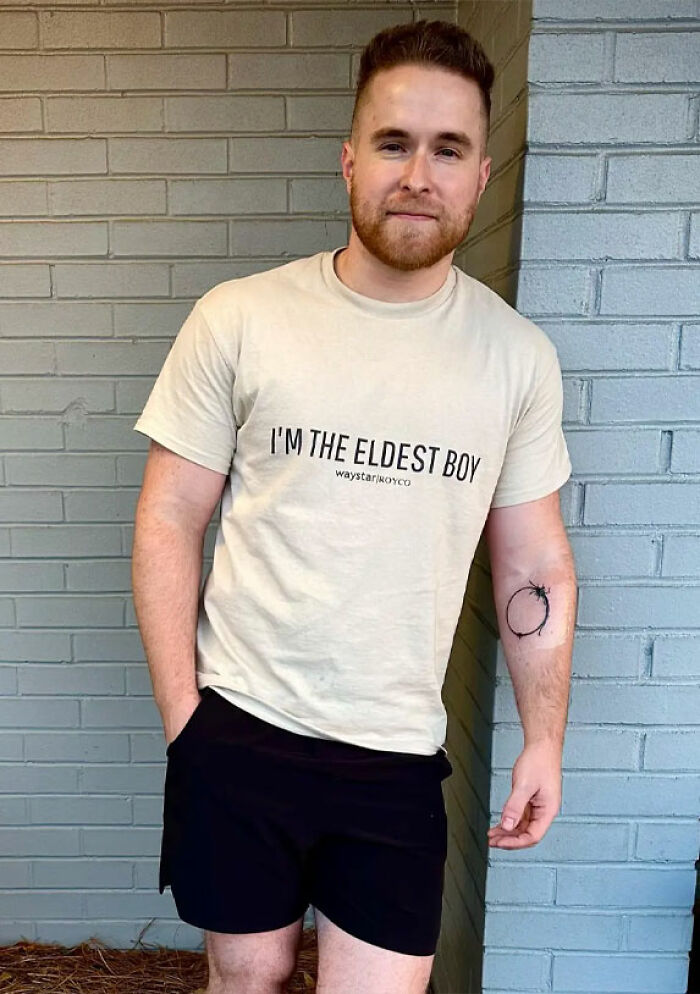 Young man standing against a brick wall wearing a beige shirt and black shorts, symbolizing short king height preferences in dating.