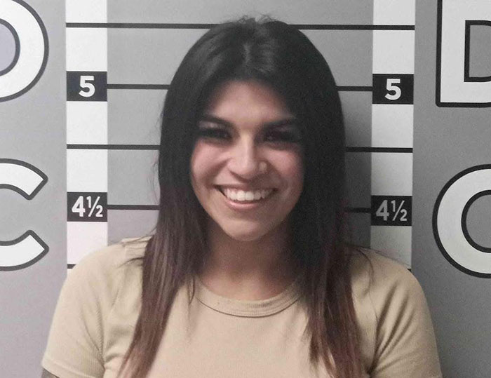 Woman in a tan shirt smiling in a mugshot photo after destroying ex&rsquo;s car by pouring salt in engine and glitter in AC.