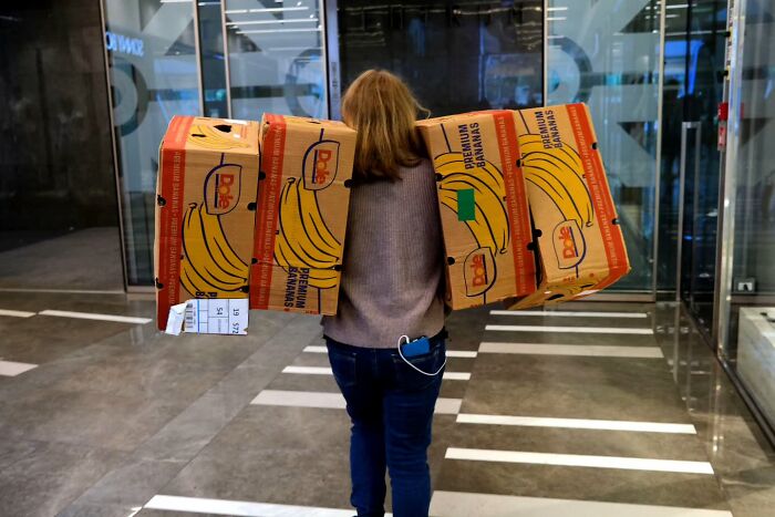 Photographer captures unseen candid street moment of a person carrying large banana boxes in an urban setting.
