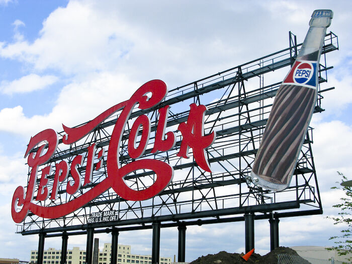 Large vintage Pepsi-Cola sign with red lettering and a retro glass Pepsi bottle against a partly cloudy sky, illustrating PR campaigns failures.
