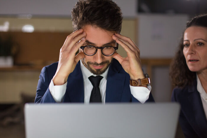 Man in glasses and suit looking stressed at laptop while woman beside him watches, reflecting modern life breaking down concerns.