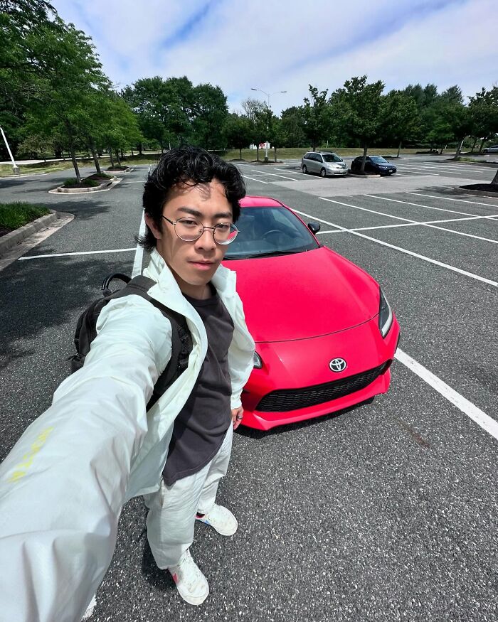 Young man taking a selfie with a red Toyota sports car in a nearly empty parking lot, highlighting celeb relatability fails.