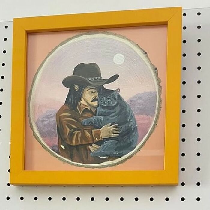 Painting of a man in a cowboy hat holding a large cat, displayed in a yellow frame at a thrift store.