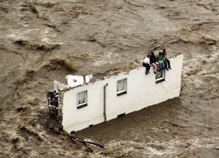 Four people sitting on a floating house wall amid floodwaters in an unsettling scene from morbid knowledge images.