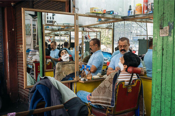 Barber shaving a customer's face in an old barbershop, capturing candid everyday moments from Asia by Gil Kreslavsky.