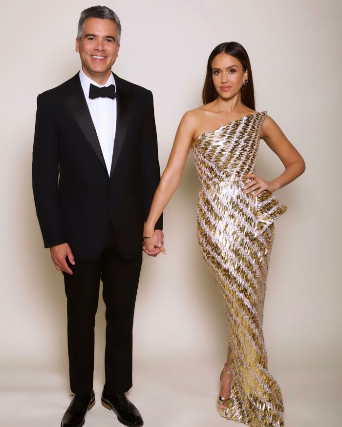 Jessica Alba posing elegantly in a metallic gown next to a man in a black tuxedo holding hands at an event.