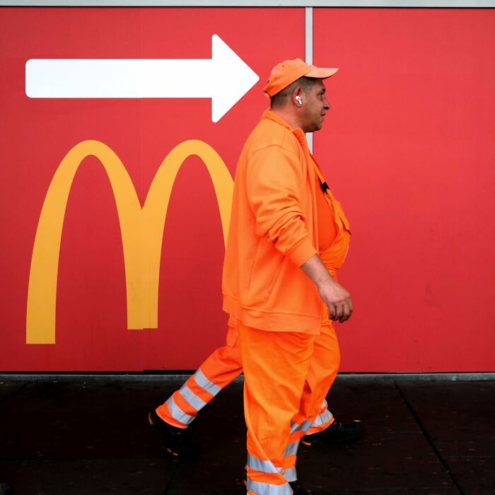 Man in bright orange workwear walking past red wall with yellow arches and white arrow in a street photographer captures unposed moments.