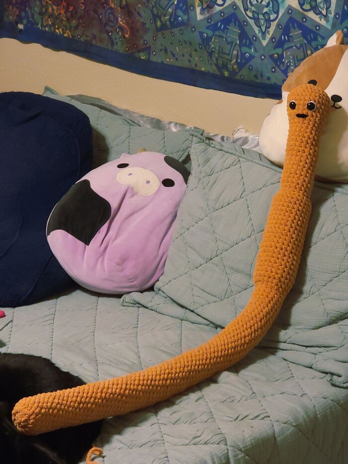 Long orange crocheted stuffed animal with googly eyes on a couch among other plush pillows, craft shaming DIY fail.