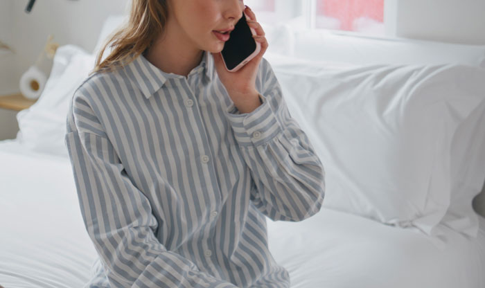 Young woman in striped shirt sitting on bed, talking on phone, dealing with family demands for money conflict.