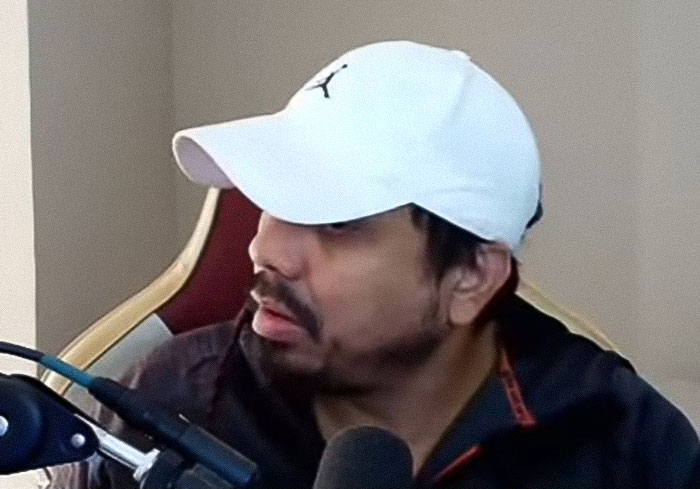 Man wearing white cap and dark jacket streaming live indoors with microphone, related to 2-year-old drowning incident.