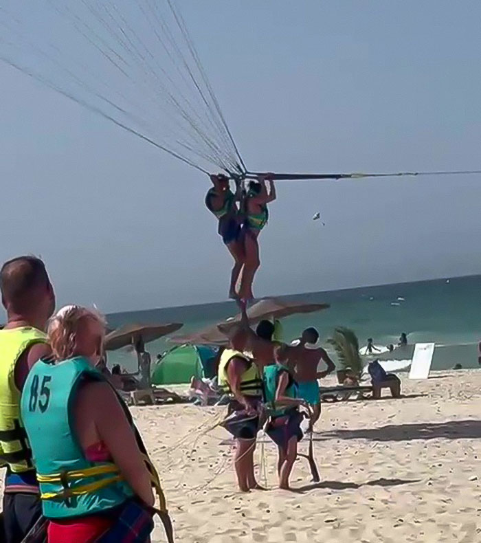 Two people parasailing mid-air above a beach with others watching, highlighting mid-air assault safety concerns. - 5