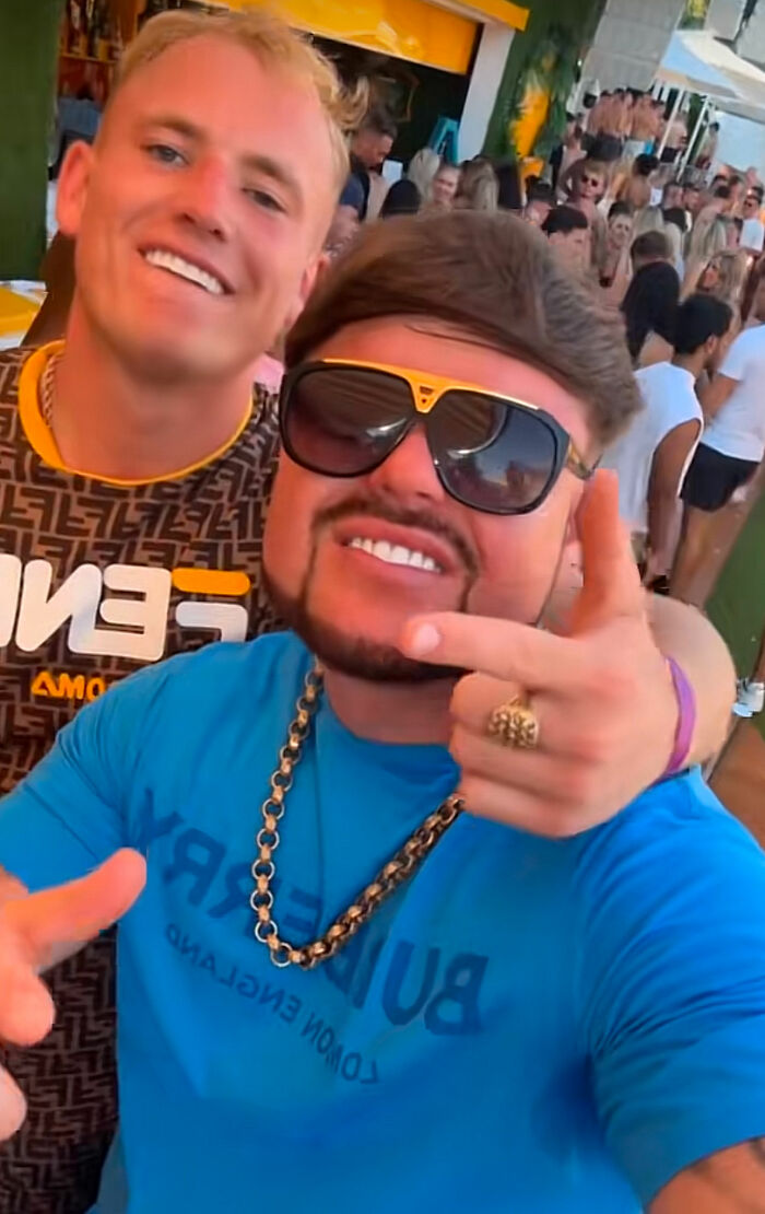 Two men posing at a crowded party, one wearing large sunglasses and a blue shirt, related to Ibiza Final Boss haircut meme frenzy. Two men posing at a crowded party, one wearing large sunglasses and a blue shirt, related to Ibiza Final Boss haircut meme frenzy.