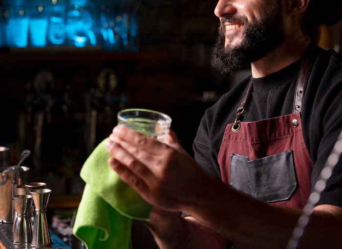 Bartender wearing apron smiling while drying glass at bar, representing bar staff protecting women from unwanted attention.