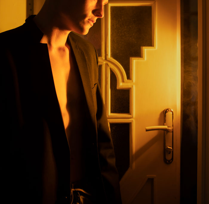 Man in a dark jacket standing near a door bathed in warm light, highlighting a mysterious and secluded atmosphere related to security camera.