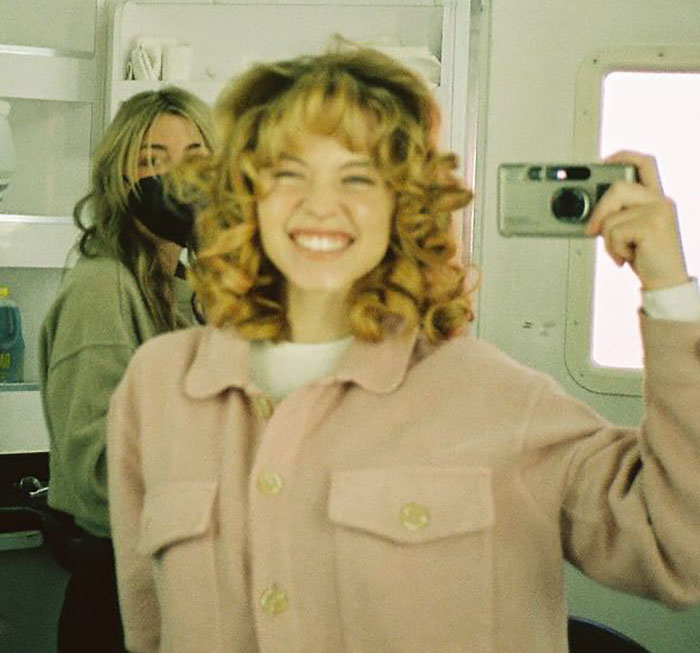Sydney Sweeney smiling and taking a selfie indoors while another person wearing a mask stands in the background.