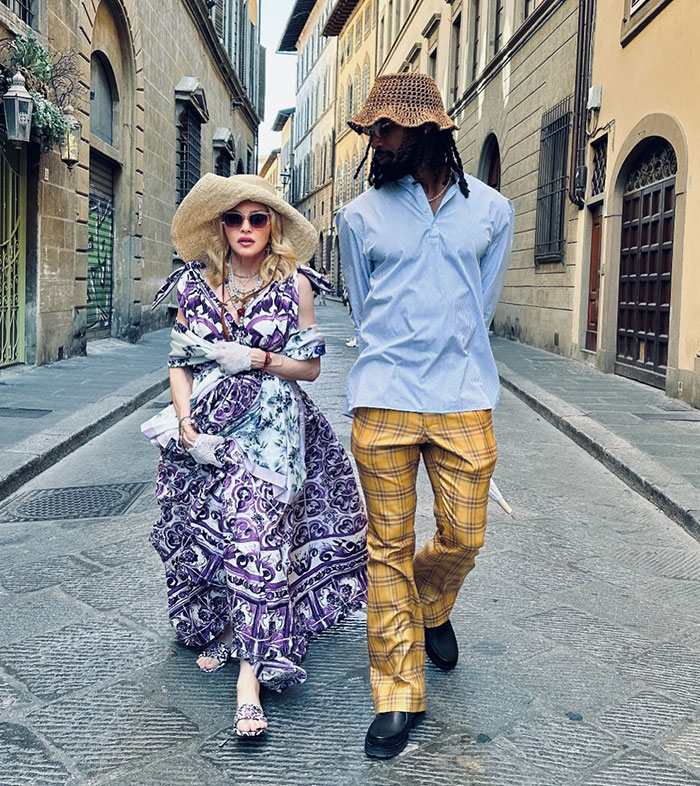 Madonna&rsquo;s daughter Lourdes wearing a patterned dress and hat, walking with a man on a cobblestone street in stylish attire.