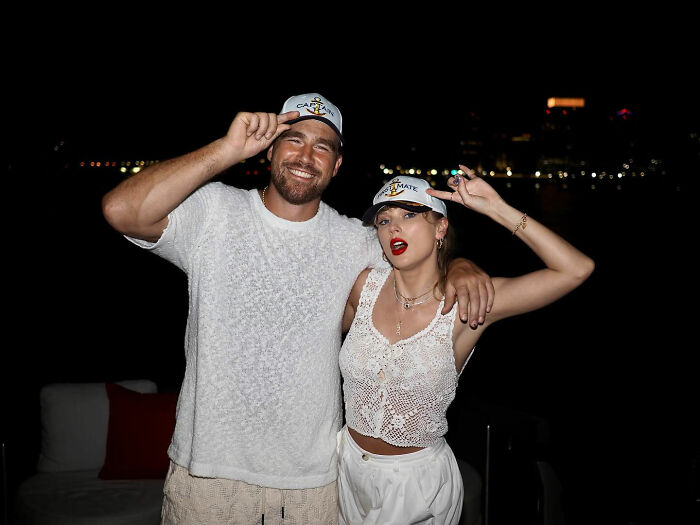 Taylor Swift and Travis Kelce posing together wearing matching caps during a nighttime event, fans debate marketing stunt.