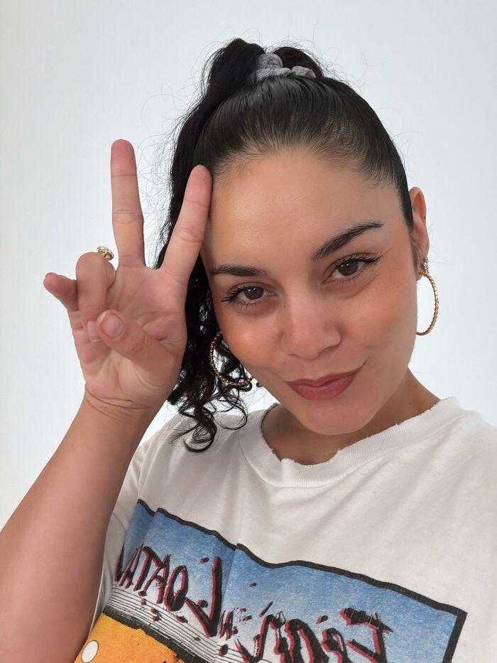 Celebrity posing with a peace sign, wearing hoop earrings and a graphic t-shirt, showing out-of-touch relatable moment.