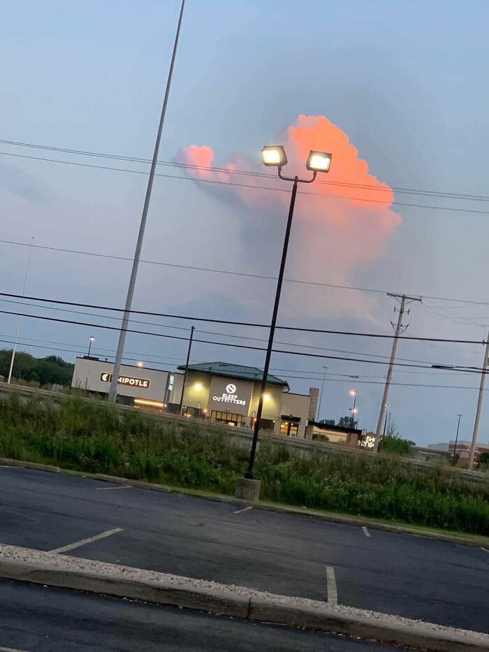 Cloud that looks like a face glowing orange at sunset behind a lit streetlamp in a parking lot, random objects resembling faces.