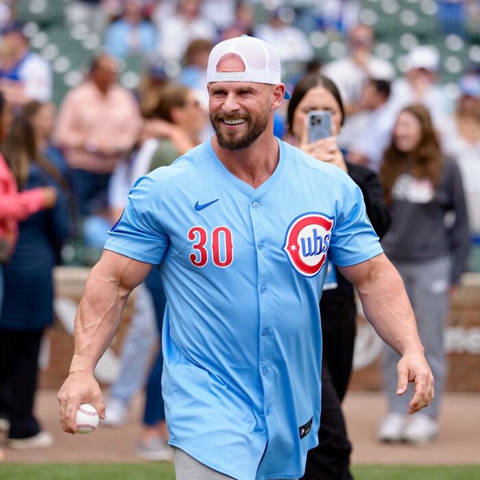Joey Swoll in a Cubs jersey smiling and holding a baseball during a public event with people in the background.