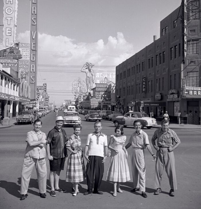Group of people posing on a busy Las Vegas street with vintage cars and historic Vegas neon signs in the background.