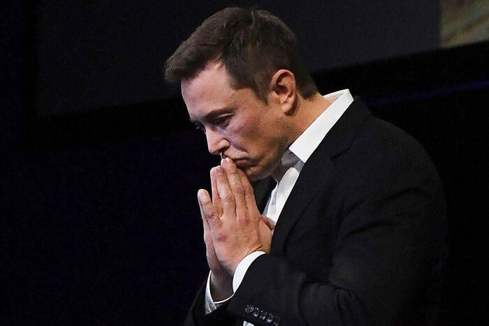 Elon Musk in a black suit, hands clasped near face, appearing thoughtful against a dark background. - 4