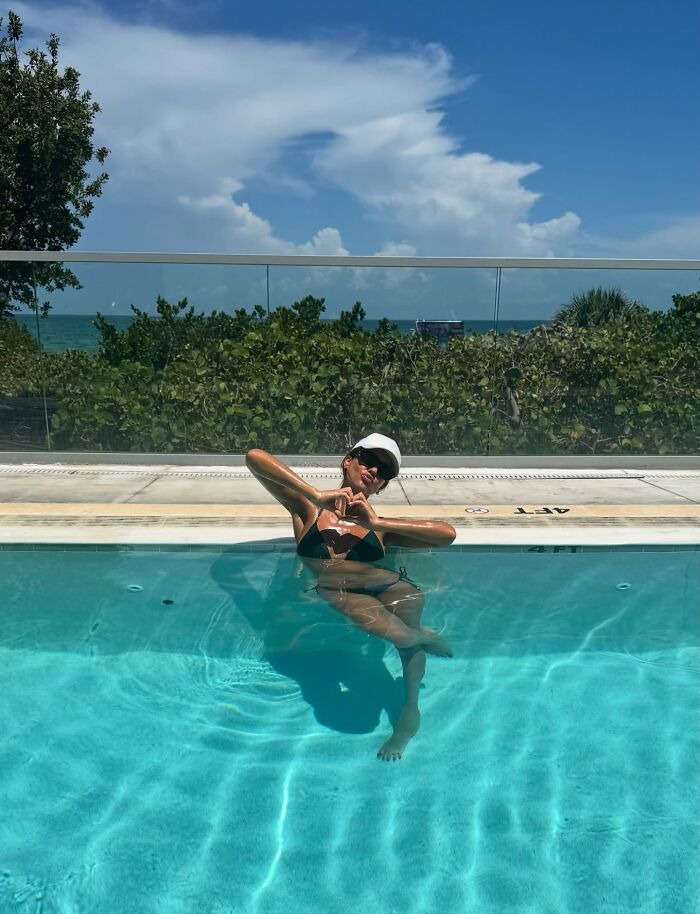 Jessica Alba posing in a black bikini and cap, relaxing in a pool with a scenic ocean and greenery backdrop.