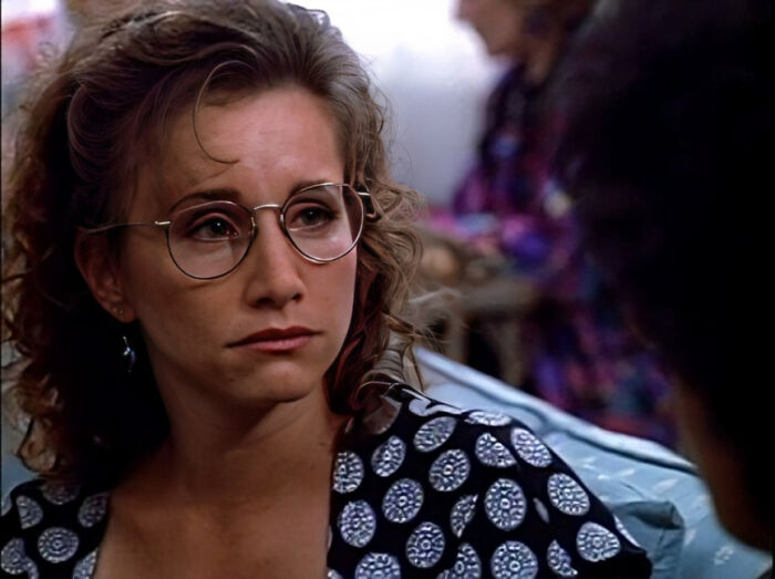 Woman with curly hair and glasses wearing a patterned dress, evoking nostalgic 90s vibes in a casual indoor setting.