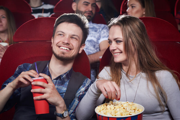 Young man and woman sharing popcorn and drinks at a movie theater, smiling and enjoying smooth pickup lines moment.