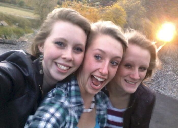 Three young women smiling and taking a selfie near train tracks with a bright light approaching in unsettling morbid knowledge image.