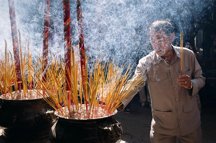 Elderly man lighting incense surrounded by smoke in a candid everyday moment from Asia captured by photographer Gil Kreslavsky