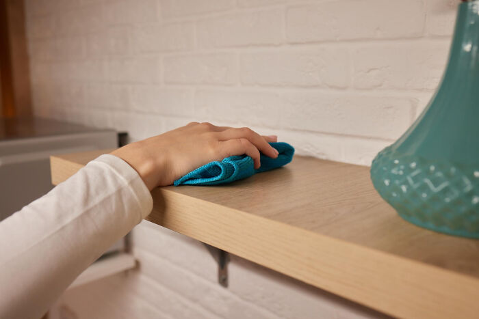 Hand wiping a wooden shelf with a blue cloth, illustrating a low effort high reward cleaning tip for easy home care.