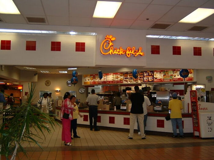 Customers waiting in line at a nostalgic 90s Chick-fil-A counter inside a mall food court with classic decor and menu boards.