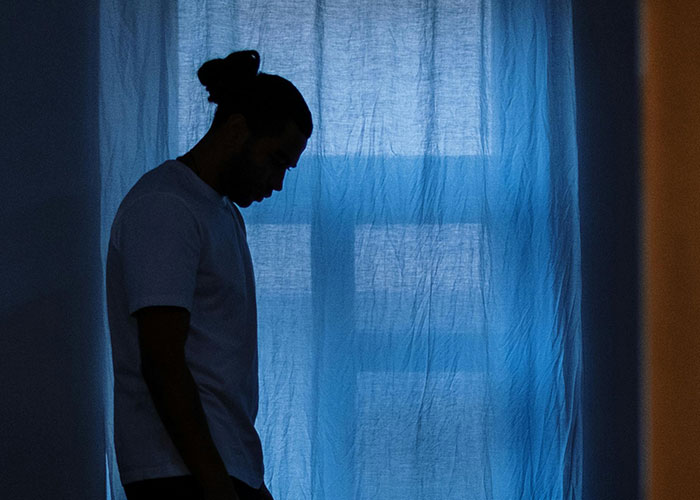 Man with hair tied back looks down in a dim room by a blue curtain, feeling sick after eating food touched by his dad.
