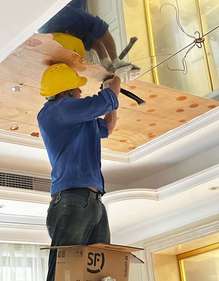 Worker wearing yellow helmet repairs broken glass ceiling and chandelier above a Labubu doll in an indoor setting.