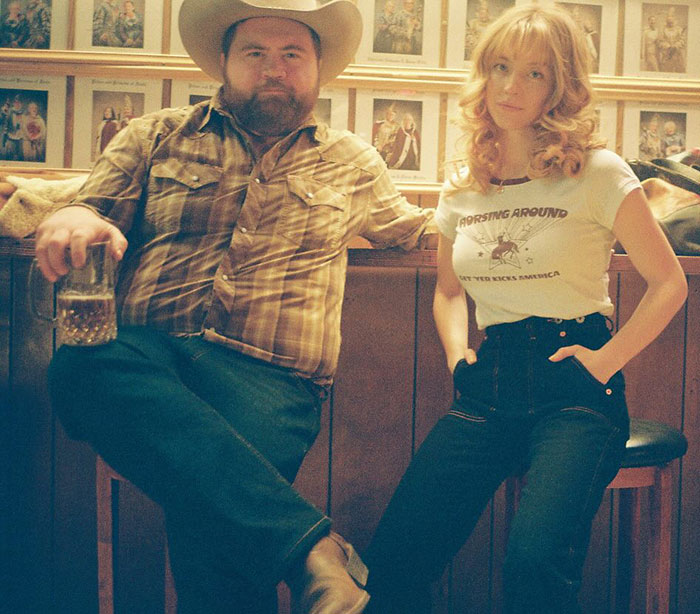 Man in cowboy hat with drink next to Sydney Sweeney wearing casual outfit in a vintage-style room setting.