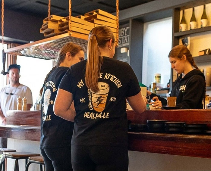 Teen caf&eacute; workers serve customers at counter in a busy coffee shop highlighting entitled customer behavior challenges.