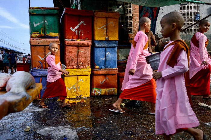 Young monks in pink and red robes walking past colorful crates in a vibrant candid everyday moment from Asia.